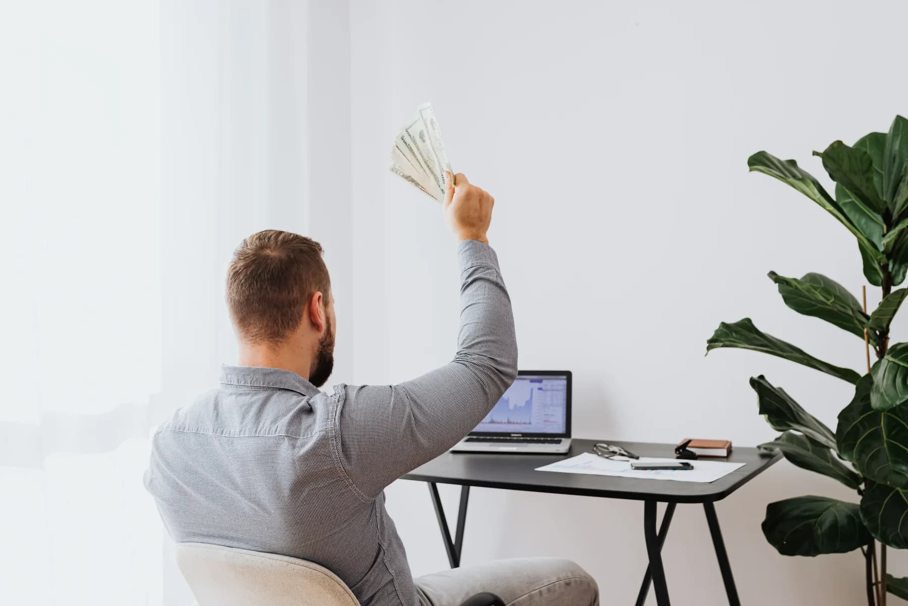 A man at a desk holding up cash in front of a laptop